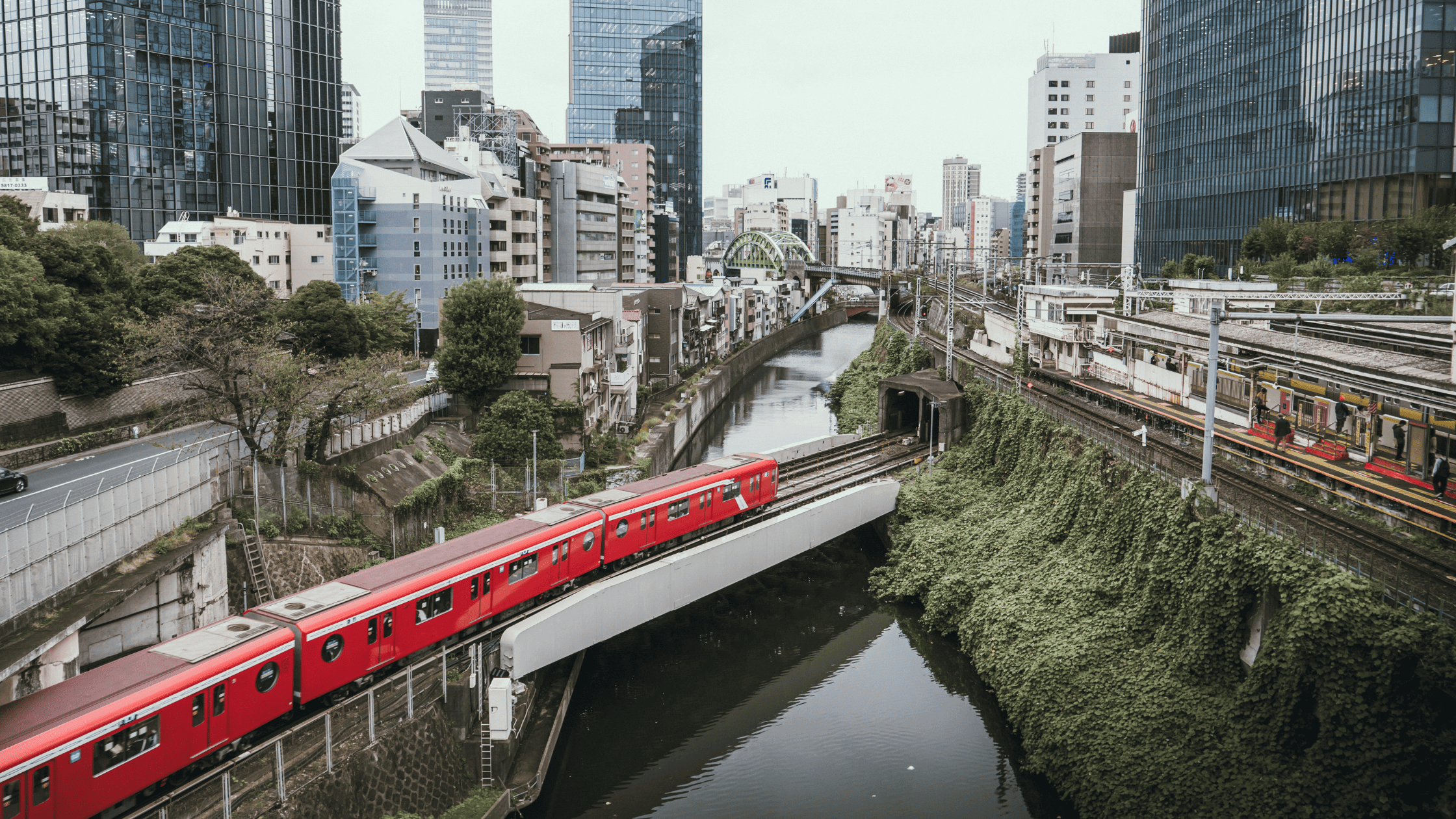 Un train rouge traversant un pont au-dessus d’un canal entouré de verdure et de bâtiments modernes à Tokyo.